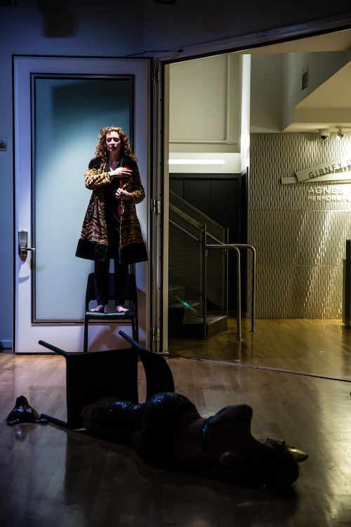 A dancer in a fur coat stands on chair holding red string as another performer lays on the ground